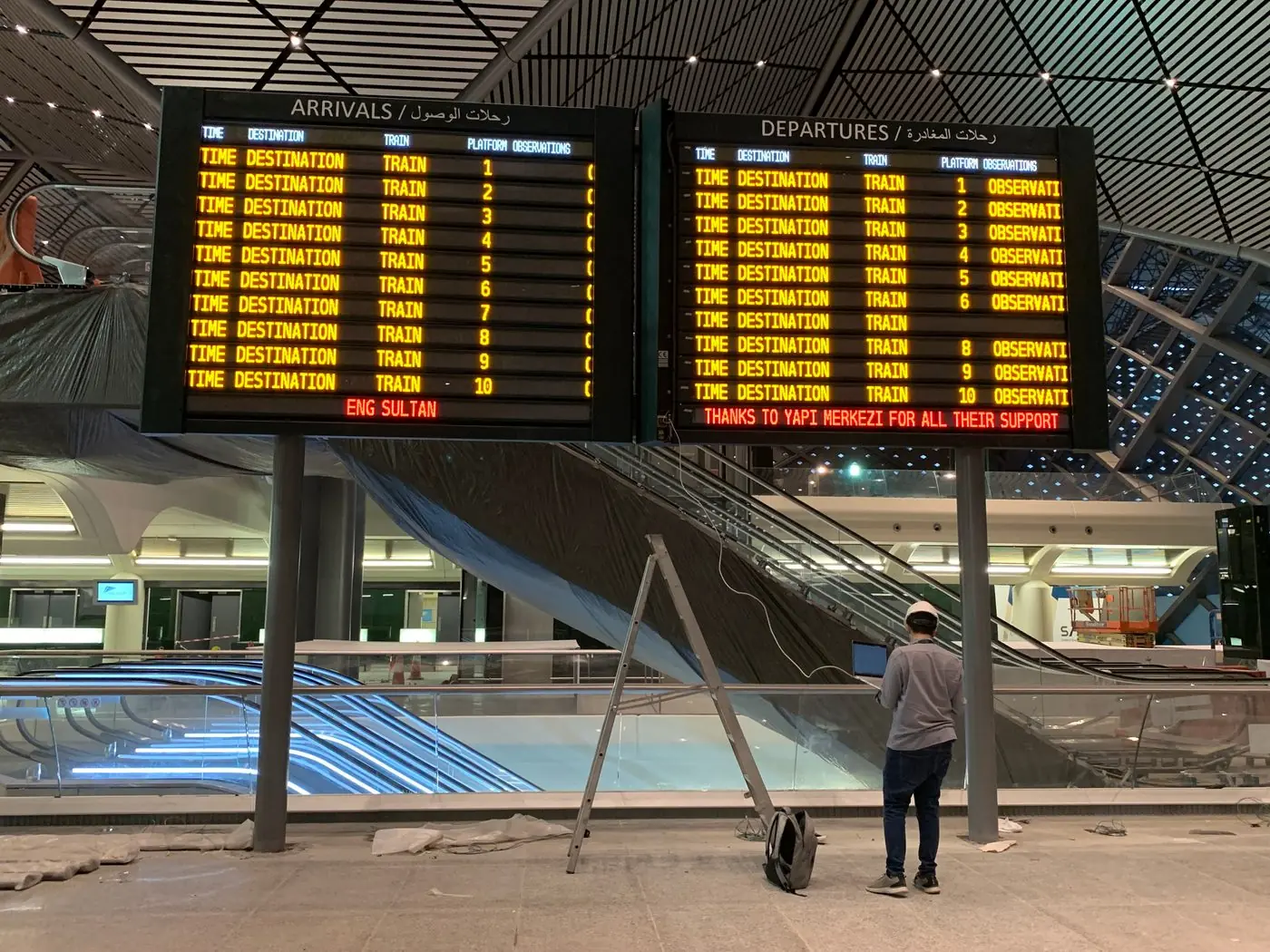 A departure board lit above a station concourse, listing services in Arabic and English.