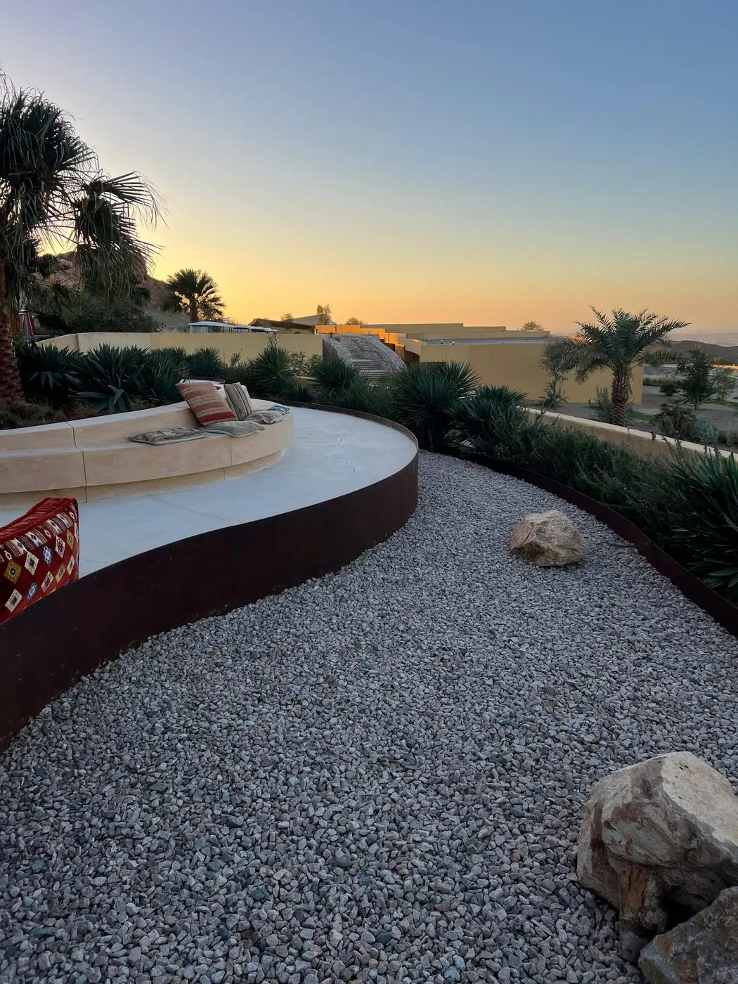 A curved Cor-Ten steel wall edging a seating terrace at dusk, with a desert resort and mountains behind it.